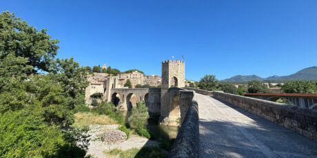 Ampia veduta del celebre Pont Vell di Besalù, il monumentale ponte medievale in pietra con la sua caratteristica torre fortificata sul fiume Fluvià, circondato da una fitta vegetazione verde sotto un cielo azzurro e limpido.