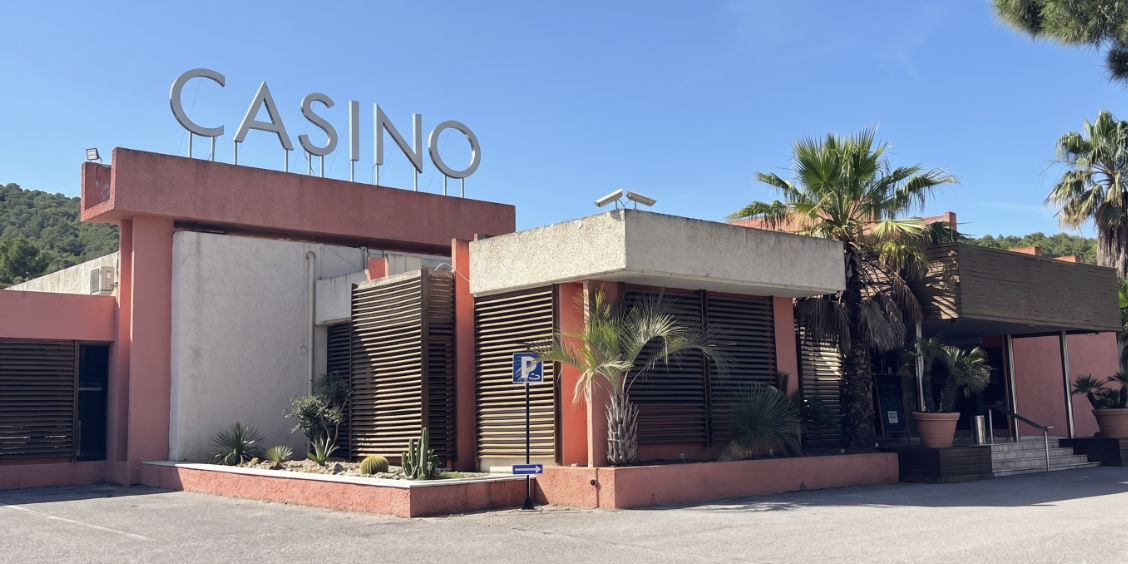 Facciata esterna del moderno Casino JOA de Le Boulou, con la grande scritta sul tetto e piante mediterranee all'ingresso.