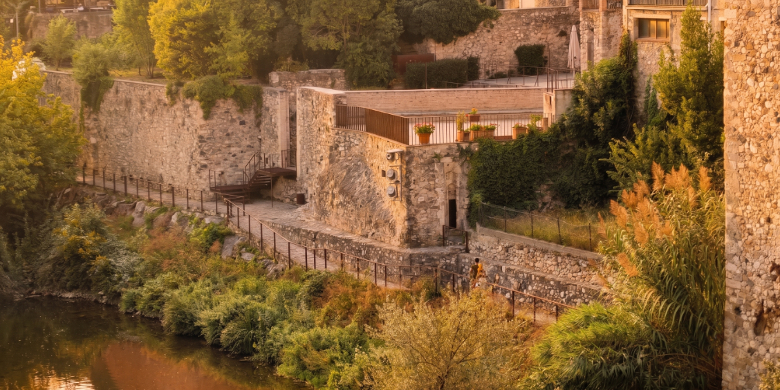 Veduta del sentiero in pietra lungo il fiume Fluvià a Besalù, sovrastato dalle storiche mura medievali del quartiere ebraico vicino al Miqvé, illuminato da una calda luce dorata al tramonto.