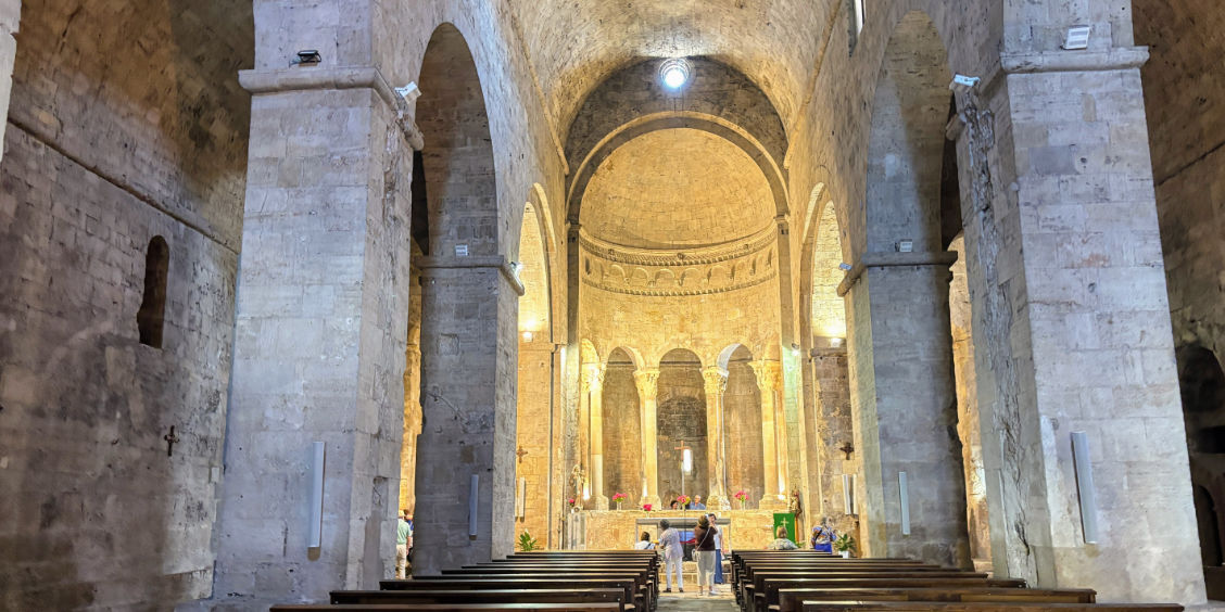 Interno in pietra del Monastero di Sant Pere a Besalù, con le sue maestose navate romaniche, i grandi pilastri ad arco e l'abside con deambulatorio a colonne dietro l'altare maggiore.
