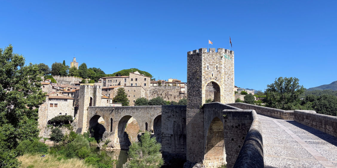Veduta panoramica diurna del celebre Pont Vell di Besalù, il monumentale ponte fortificato medievale in pietra con le sue arcate asimmetriche sul fiume Fluvià e la torre difensiva con bandiere sotto un cielo azzurro e limpido.