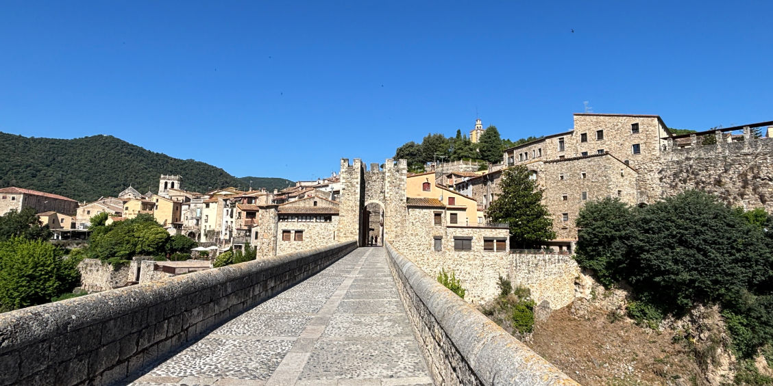 Prospettiva dal selciato in pietra del Pont Vell di Besalù rivolta verso la torre d'ingresso fortificata del borgo medievale, con le case in pietra a ridosso del fiume e un cielo azzurro limpido.